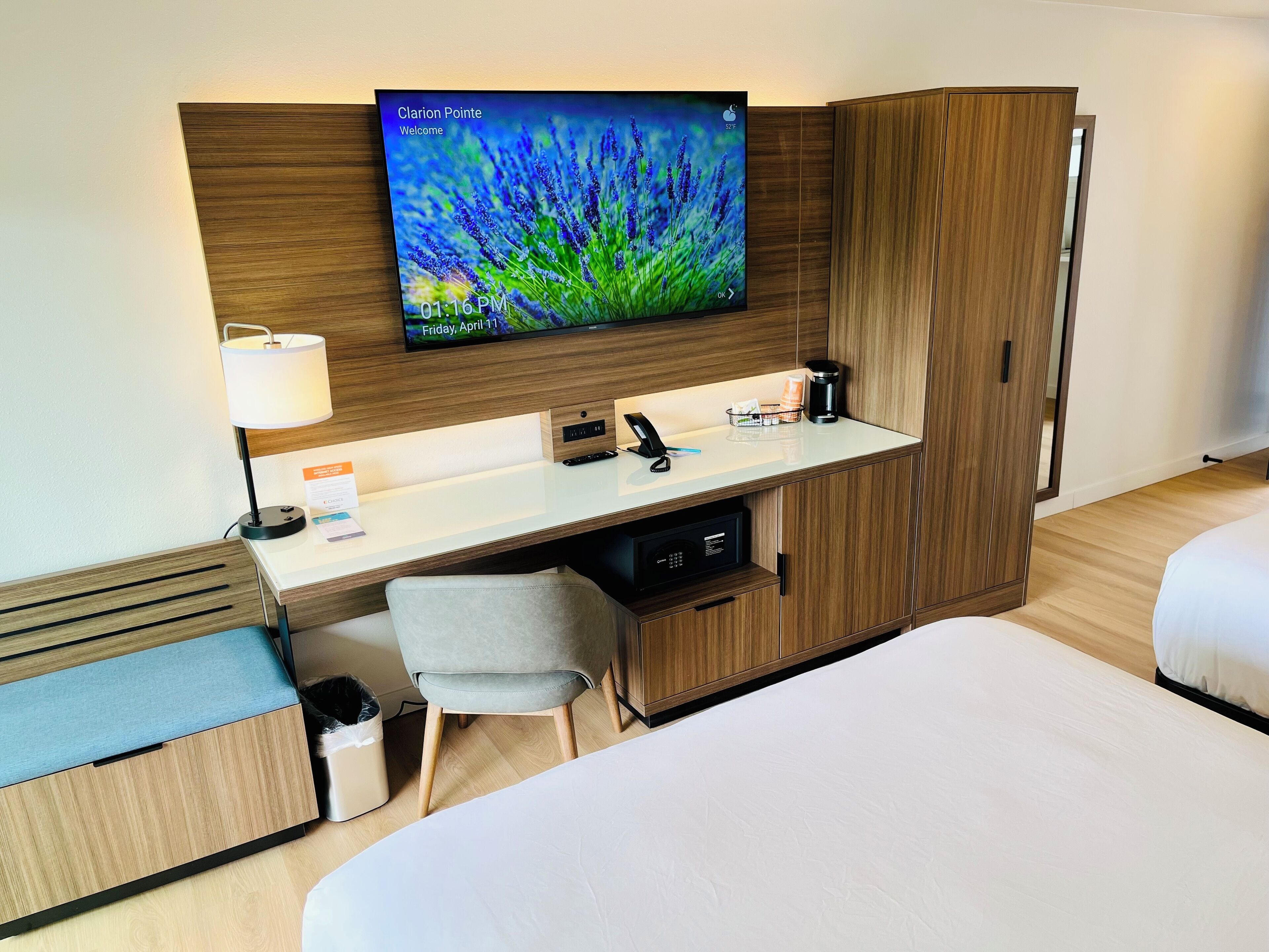 Habitación de hotel con una cama, un escritorio con monitor de computadora, una lámpara, una cafetera y un cubo de basura.