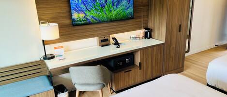 A hotel room with a bed, a desk with a computer monitor, a lamp, a coffee maker, and a trash bin.