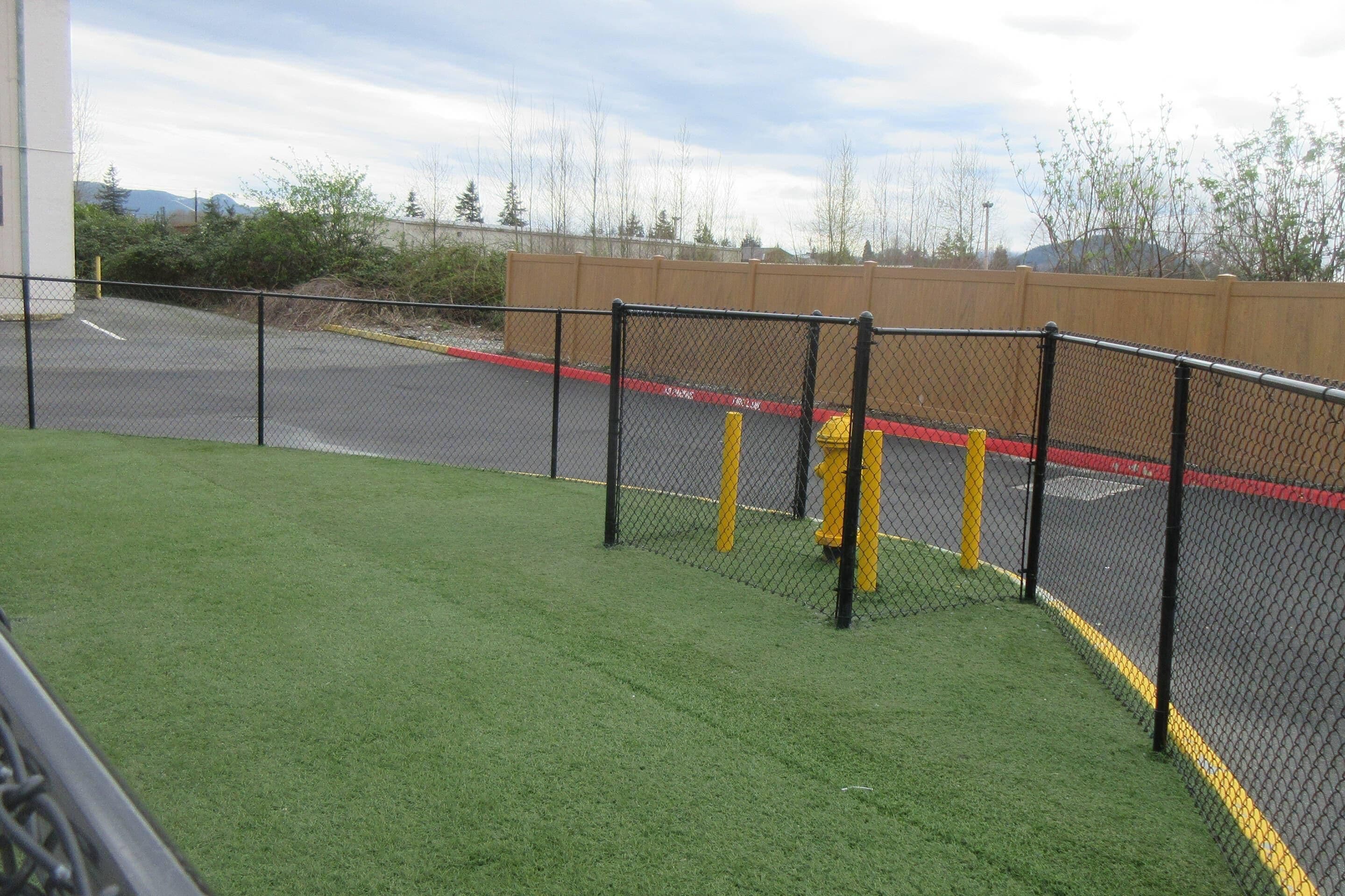 Un área de estacionamiento cercada con césped artificial verde al frente, una valla de madera y un bolardo amarillo.