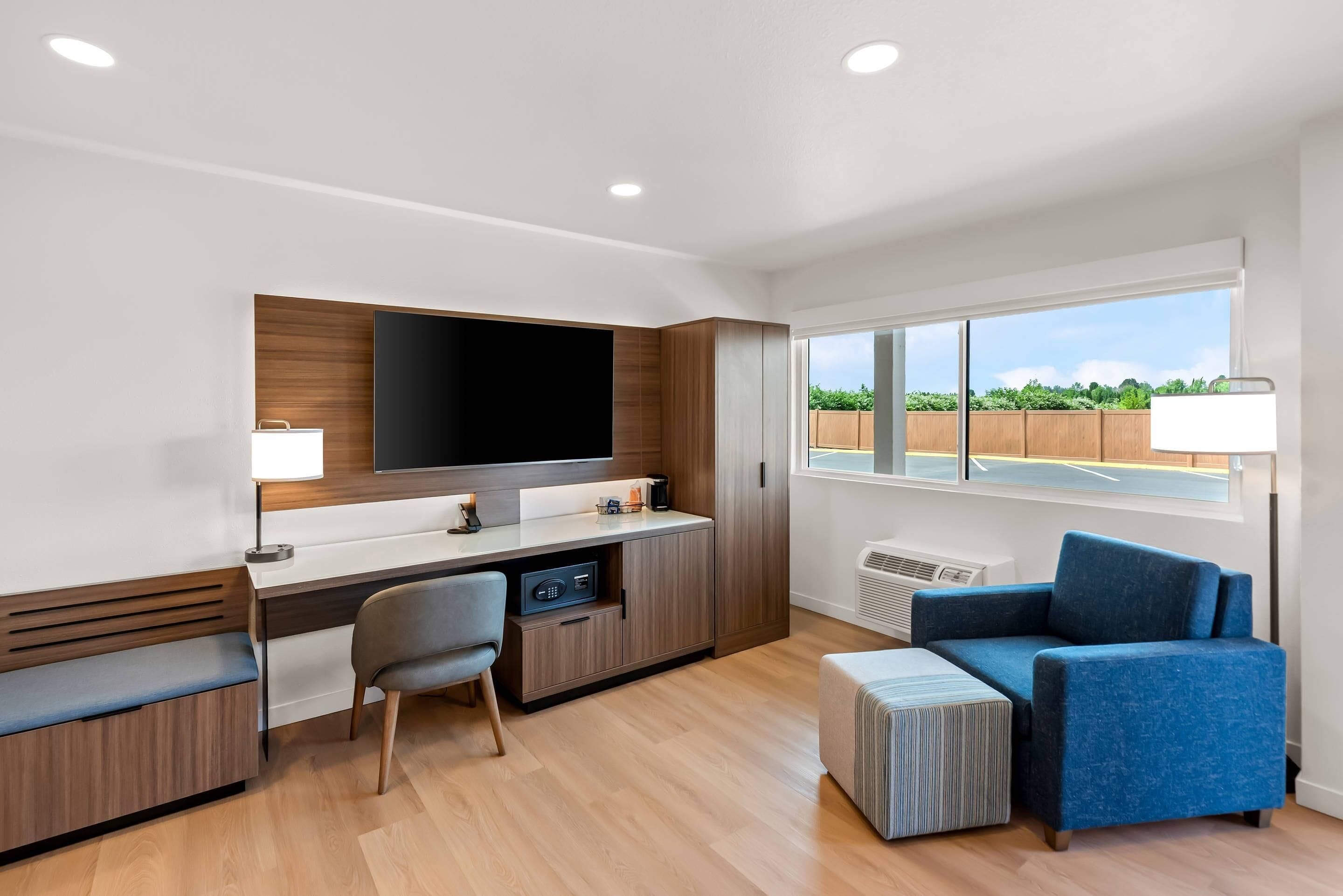 A modern living room with a flat-screen TV, a desk, a chair, a blue sofa, and a large window with a view of greenery.