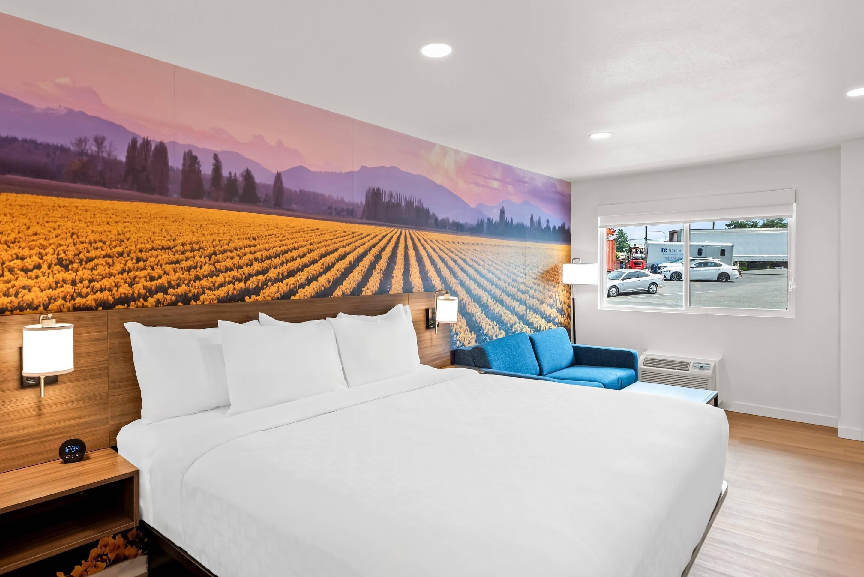 A hotel room with a large bed, a blue chair, a nightstand with a lamp, and a mural of a field and mountains.