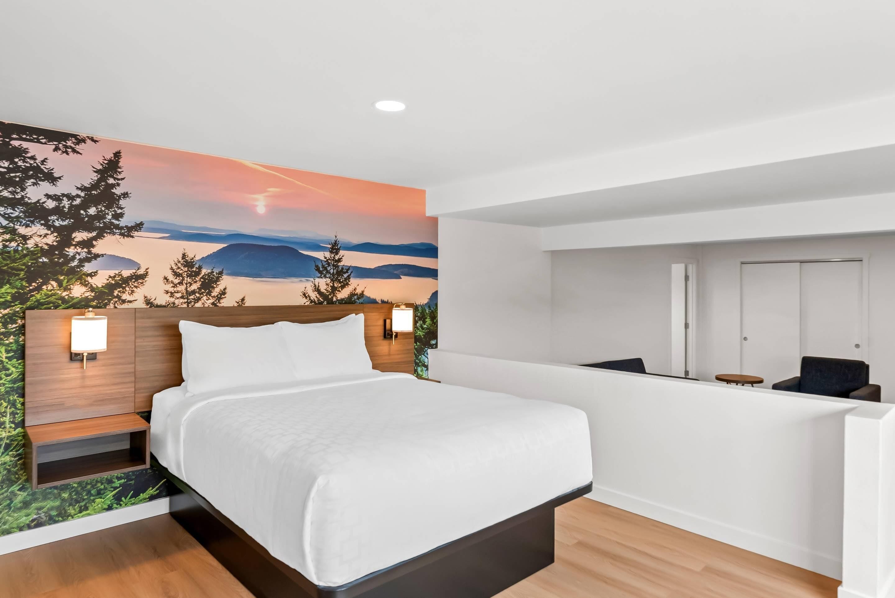 A modern hotel room with a bed, a desk, and a view of a sunset landscape.