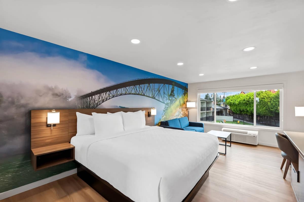 A modern hotel room with a large bed, a nightstand, a desk, and a view of a bridge through a window.
