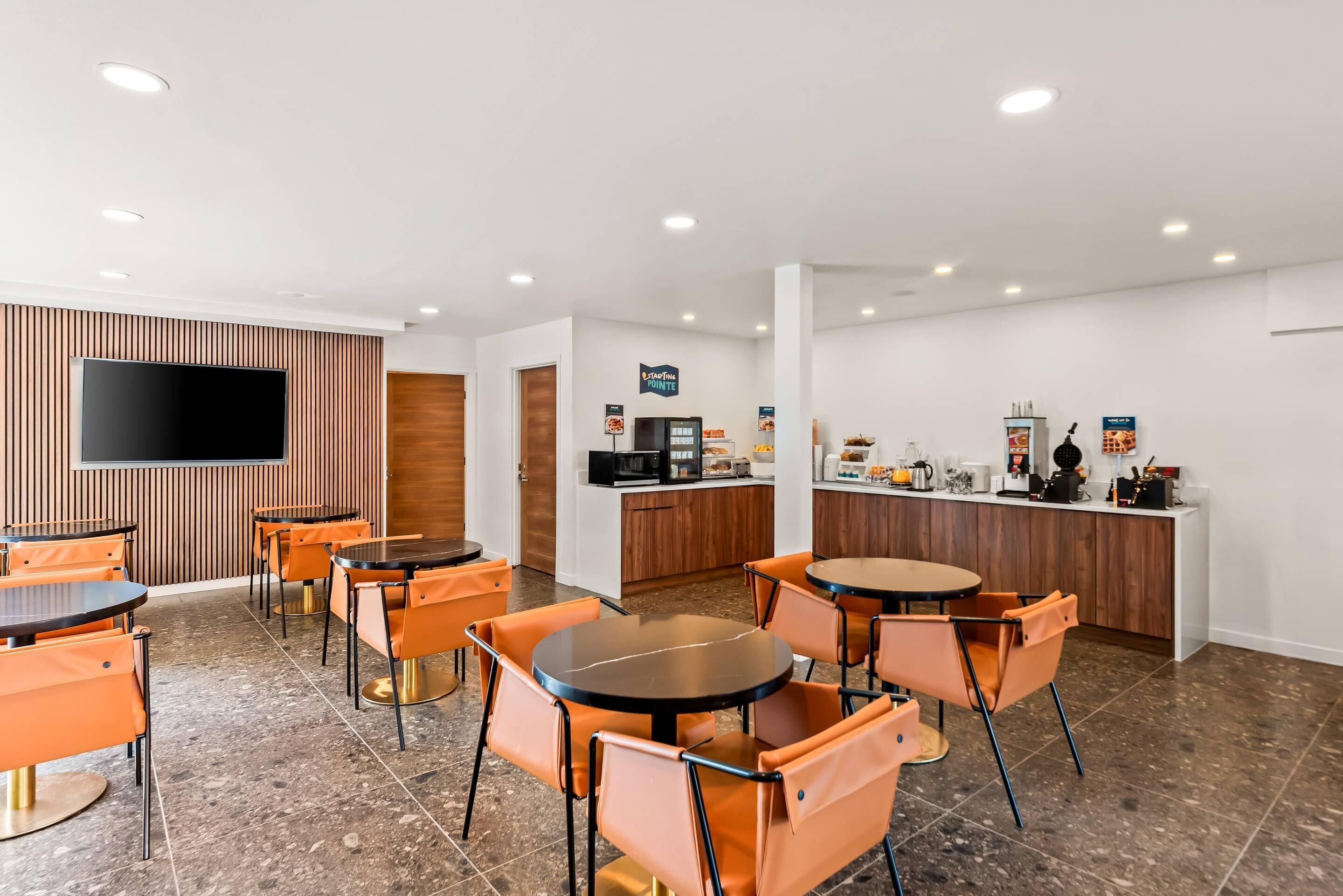 Um hall de hotel moderno com uma televisão de tela plana fixada em uma parede de painéis de madeira, uma área de kitchenette com máquina de café, e várias cadeiras e mesas laranjas dispostas para refeições.