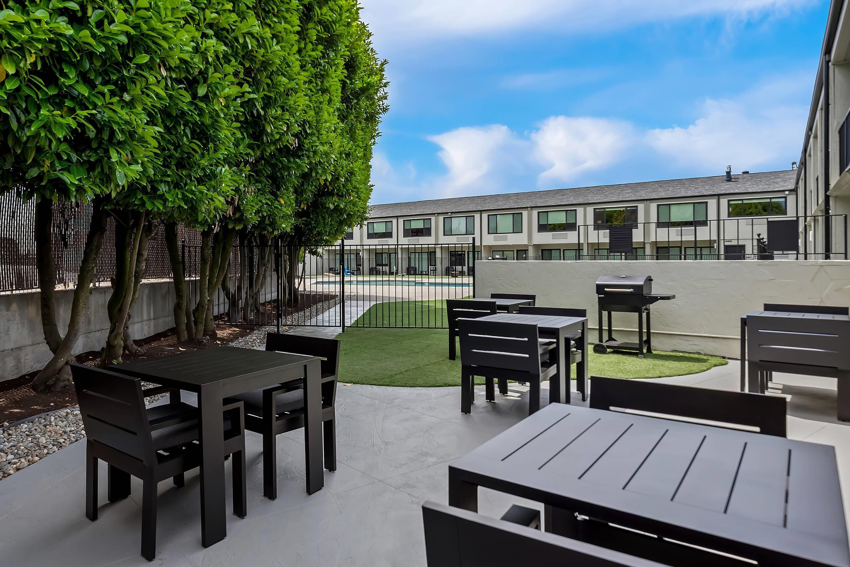 An outdoor patio area with tables and chairs, a small lawn, and a building in the background.