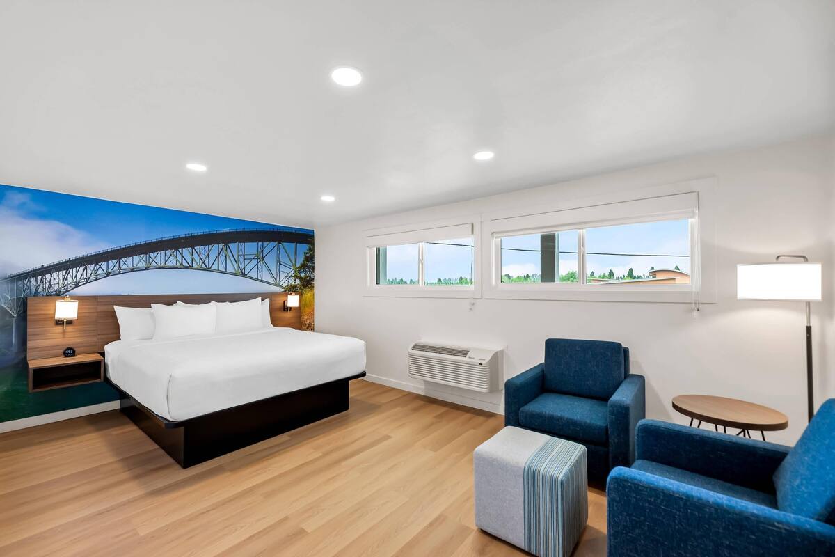 A modern hotel room with a large bed, a blue armchair, a small table, and a mural of a bridge.
