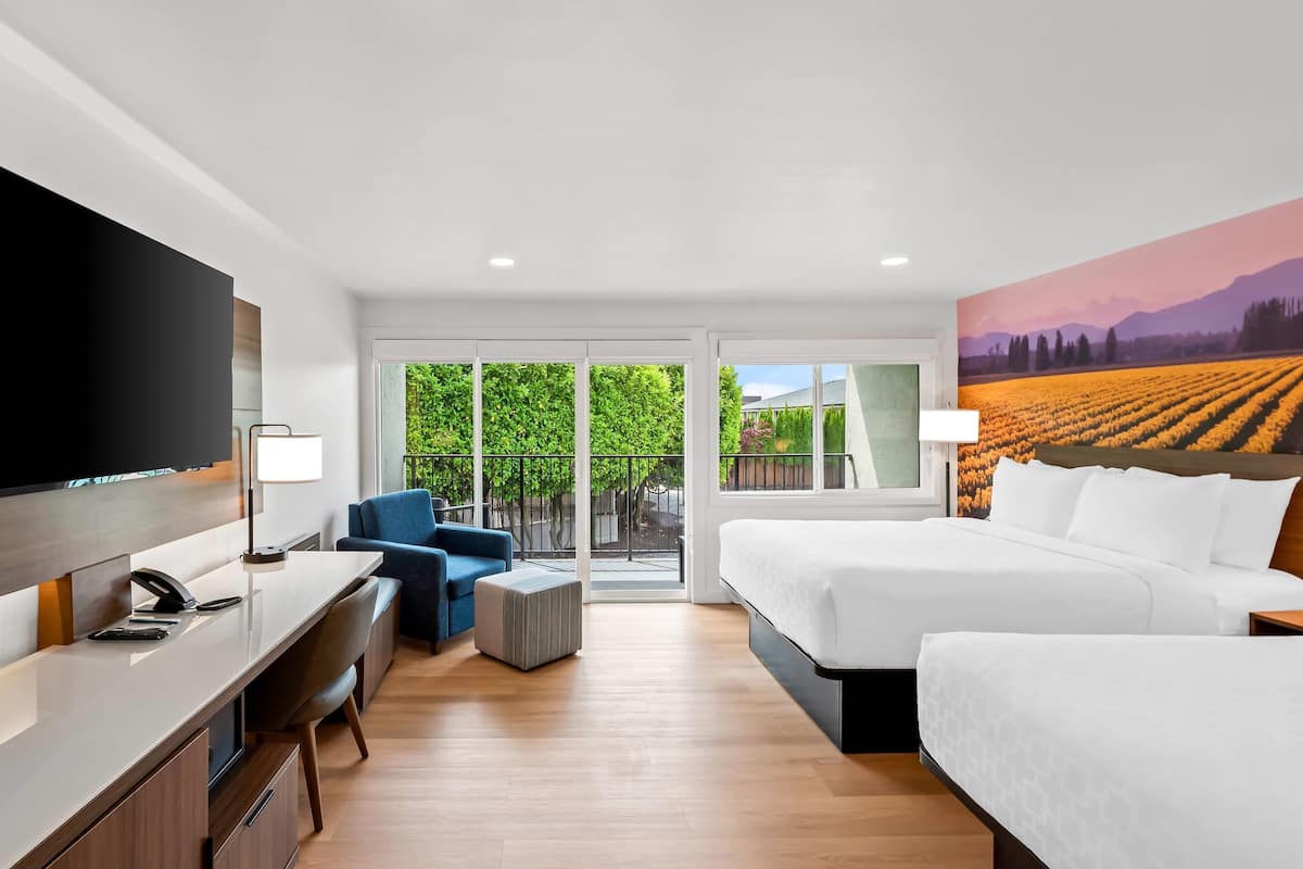 A hotel room with a large bed, a desk with a chair, a TV, and a balcony with a view of a field.