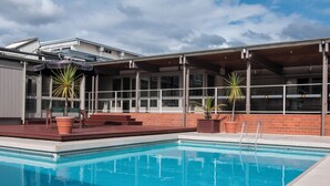 A swimming pool with a wooden deck, potted plants, and a covered area with large windows. - Copthorne Hotel Rotorua (Rotorua)