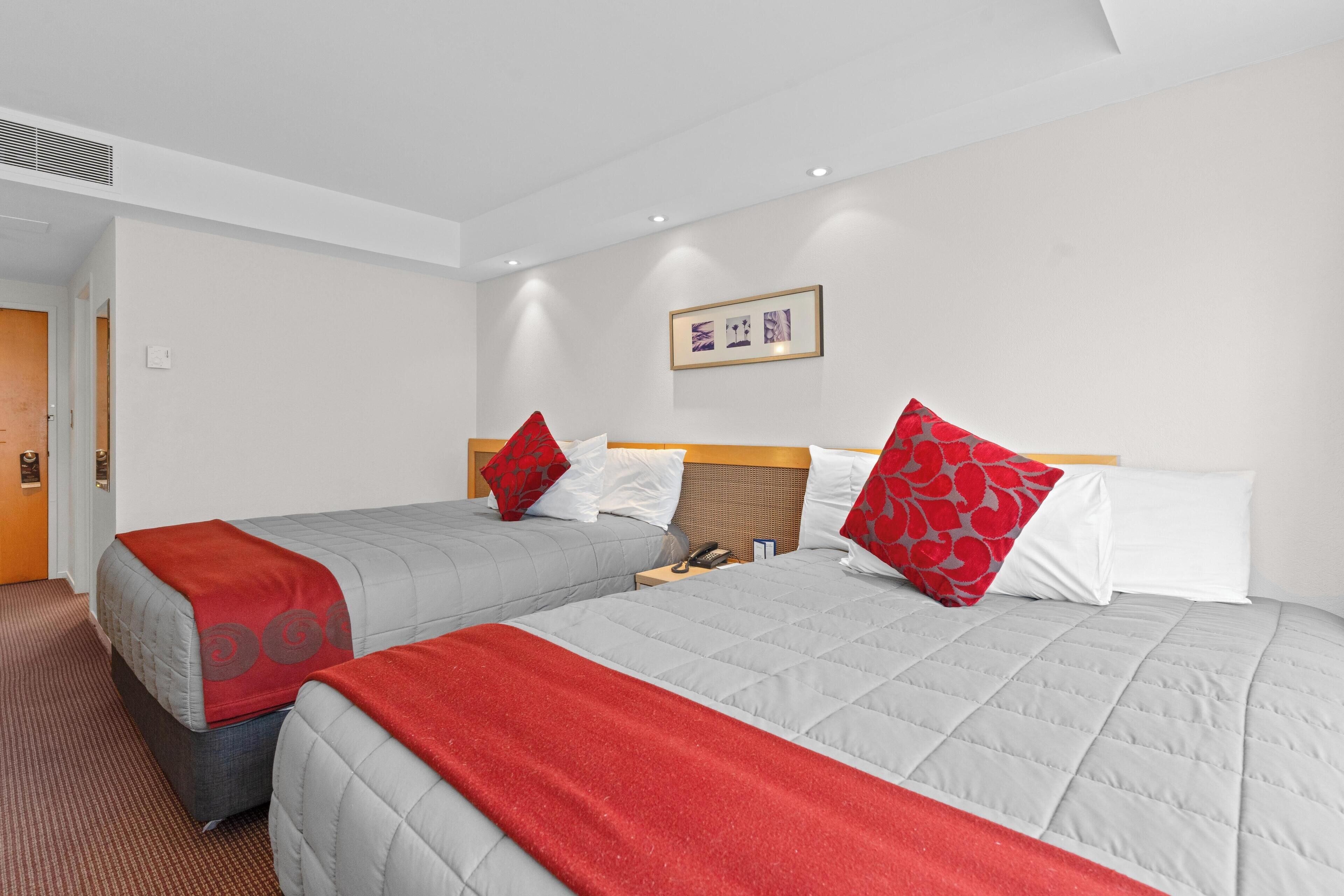 A hotel room with two beds, each with a red and grey blanket, and a wooden headboard. There is a framed picture on the wall and a door to the left.