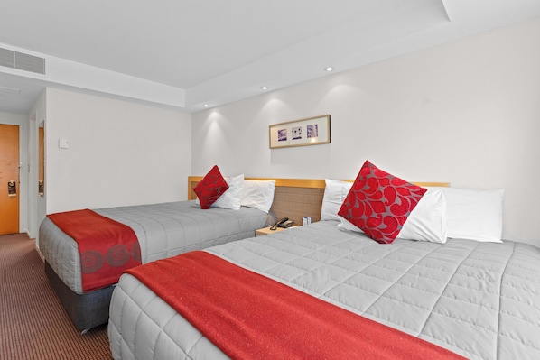 Habitación de hotel con dos camas, cada una con una manta roja y gris, y un cabecero de madera. Hay un cuadro en la pared y una puerta a la izquierda.