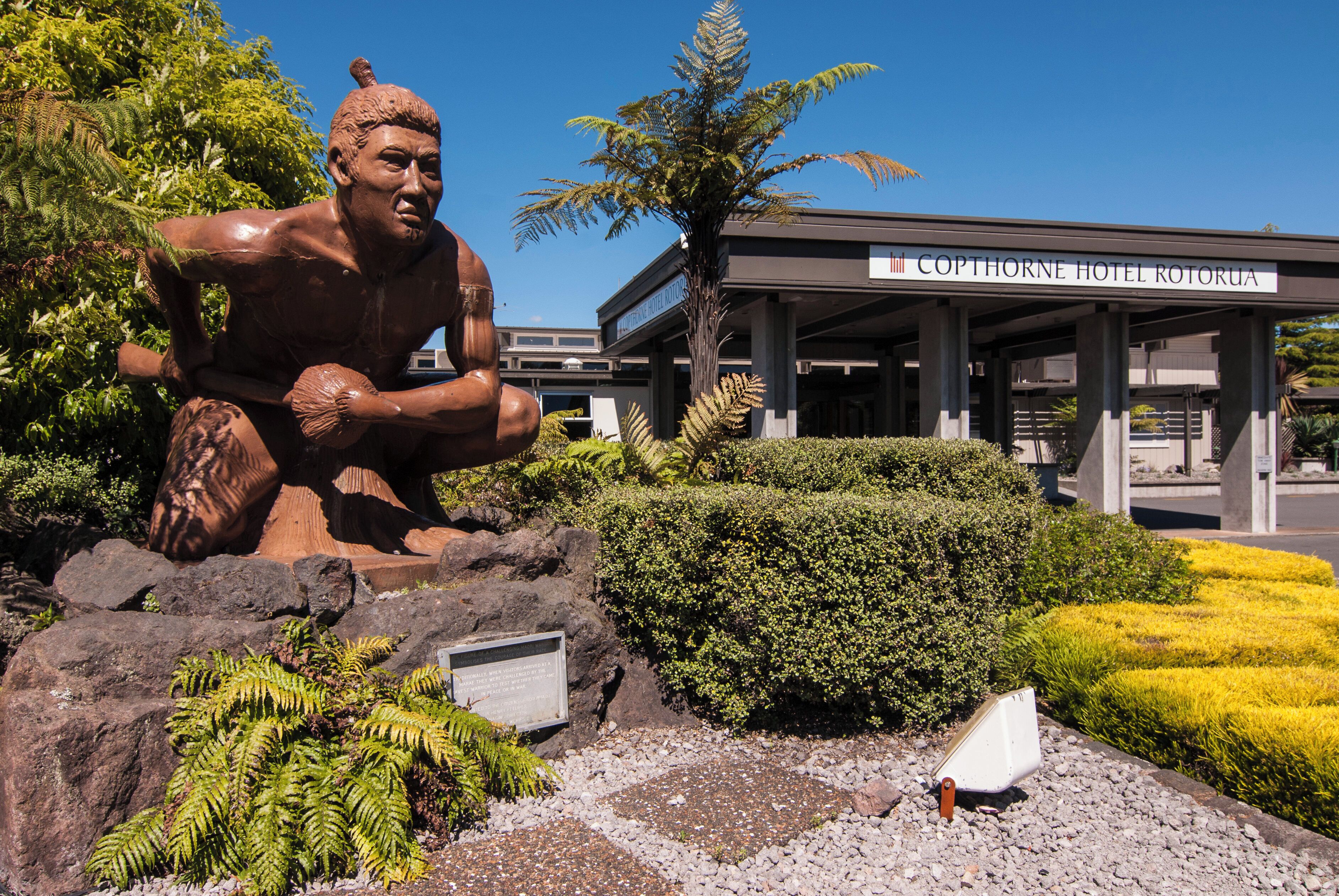 A statue of a muscular figure in a dynamic pose, a well-maintained garden with ferns and yellow flowers, and the Copthorne Hotel Rotomaru building in the background.