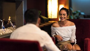 A couple seated at a table with a glass of wine, a decorative fire feature, and a red upholstered chair. - Copthorne Hotel Rotorua (Rotorua)