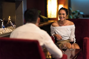 A couple seated at a table with a glass of wine, a decorative fire feature, and a red upholstered chair.