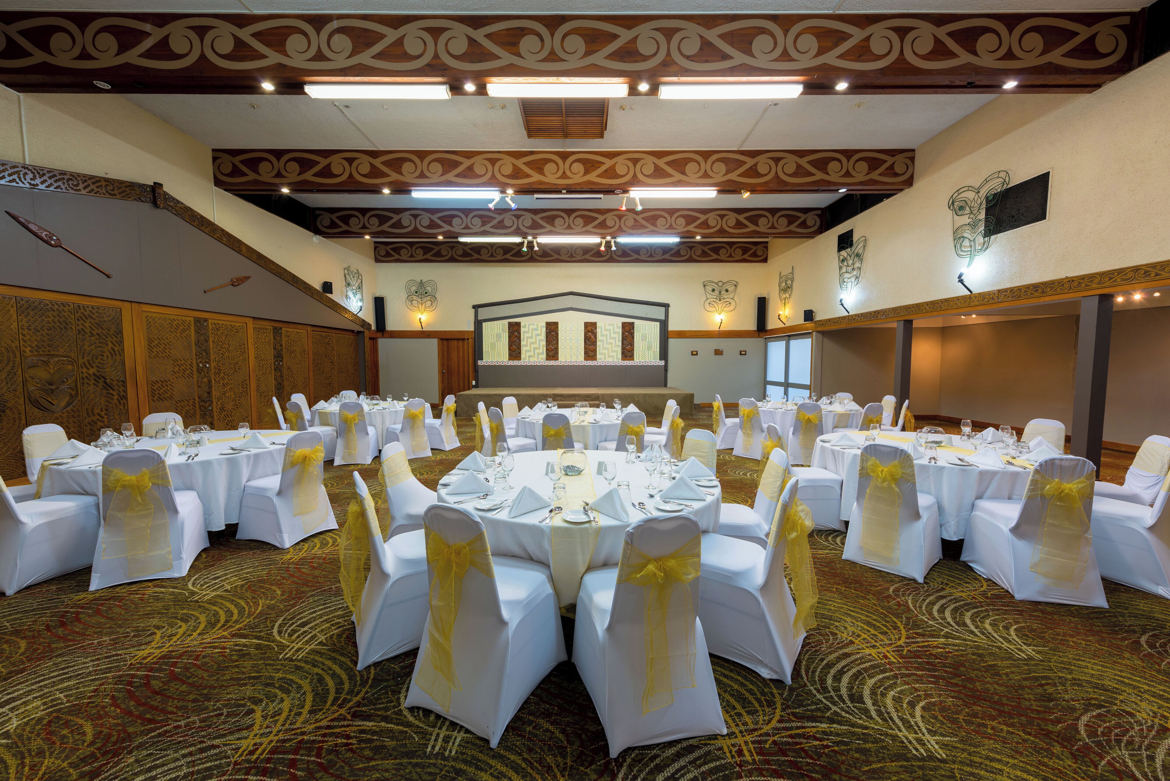 A banquet hall with round tables, white chairs with yellow sashes, and a decorative ceiling.