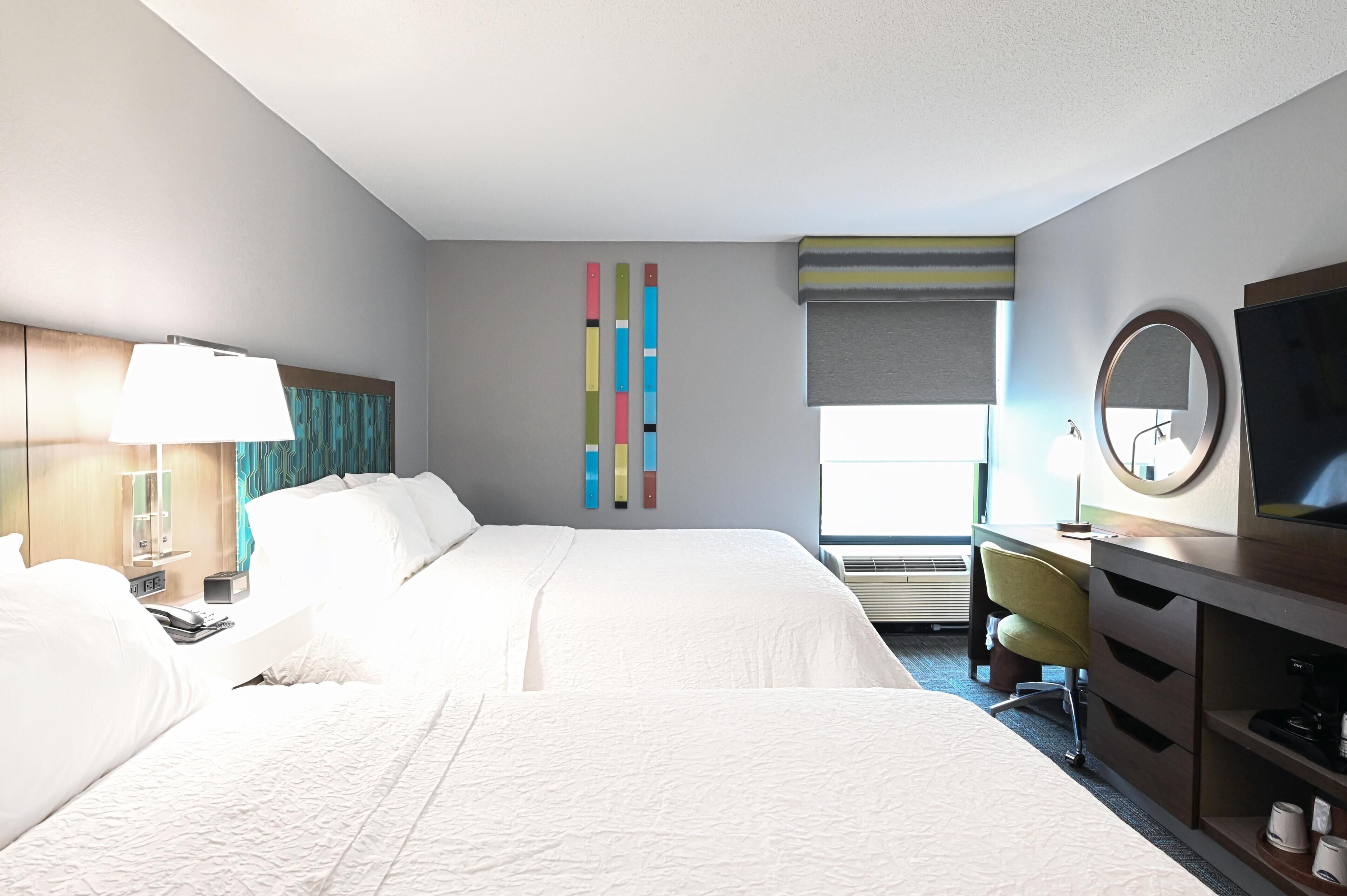 A hotel room with a large bed, a desk with a chair, a TV, a lamp, and a window with blinds.