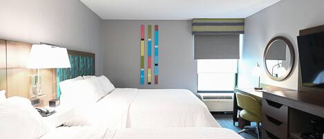 A hotel room with a large bed, a desk with a chair, a TV, a lamp, and a window with blinds.