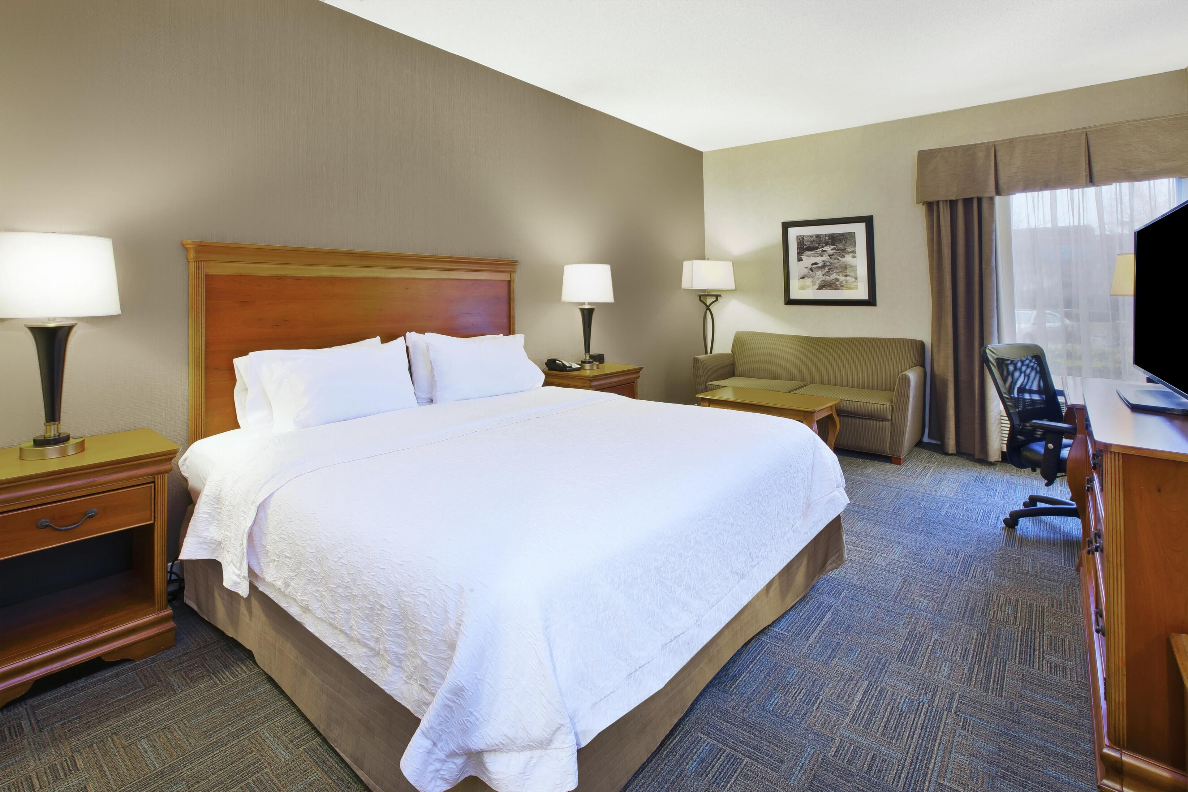 A hotel room with a large bed, two bedside tables with lamps, a desk with a chair, a sofa, and a television.