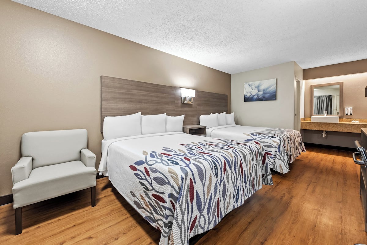 Une chambre d’hôtel avec deux lits, un canapé, un plancher en bois, une salle de bain avec un lavabo et un miroir, et un tableau accroché au mur.