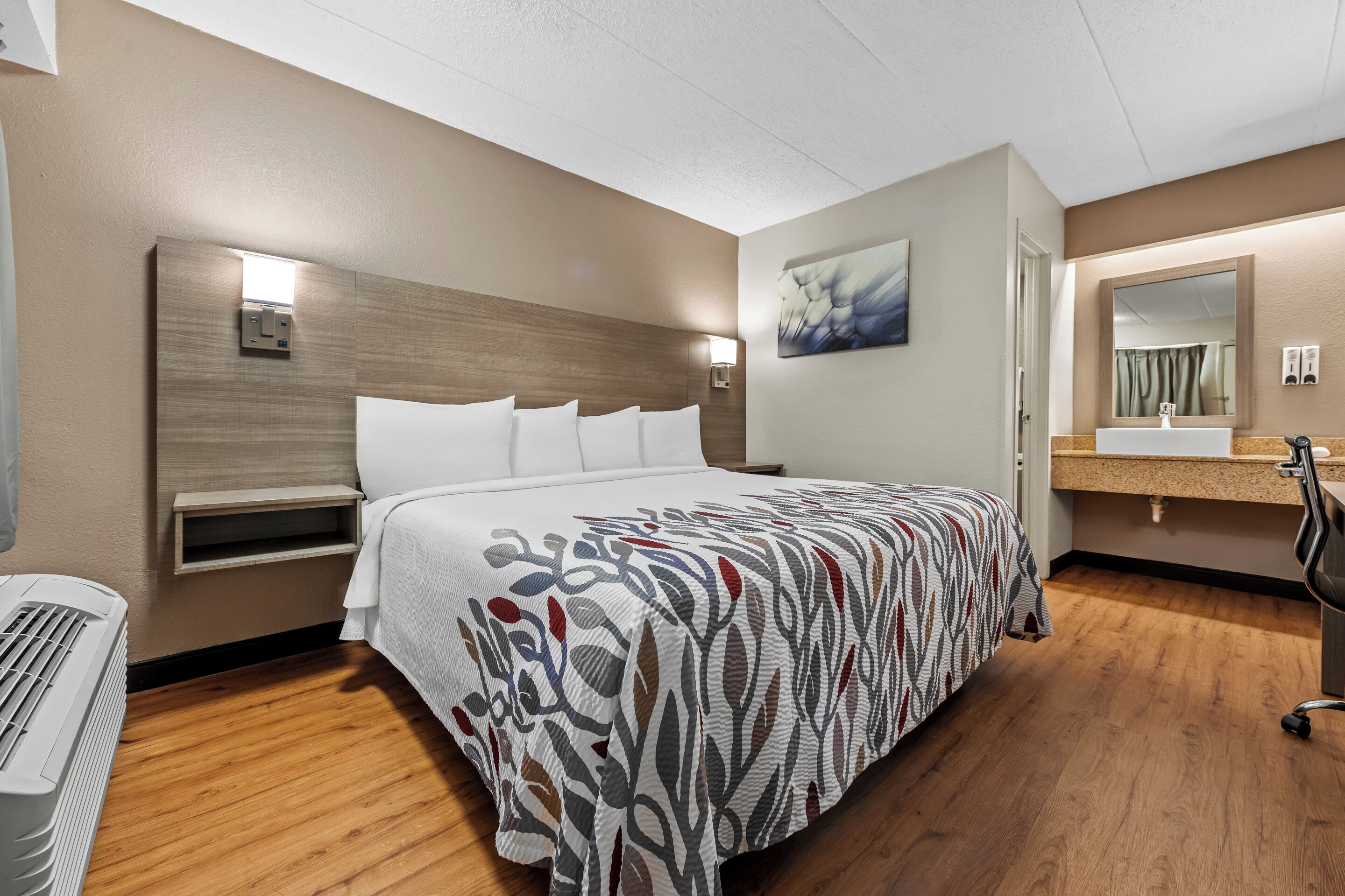 Una habitación de hotel con una cama grande, una mesita de noche, un calentador de pared, un espejo de pared, un tocador y una zona de ducha.