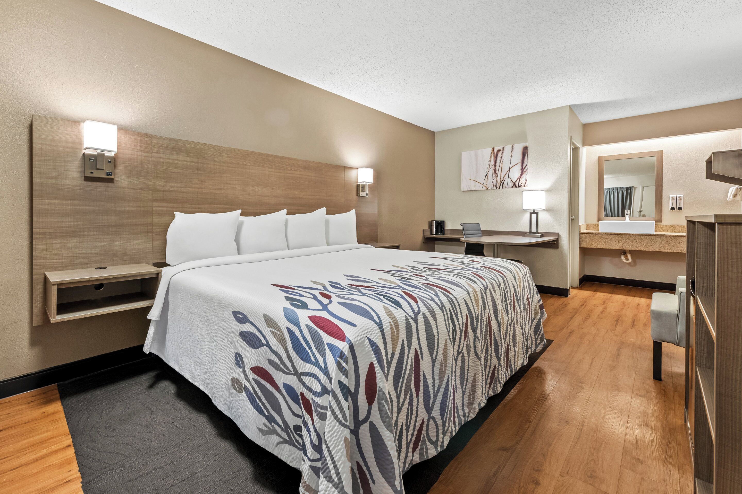 Una habitación de hotel con cama, mesita de noche, lámpara y baño con lavabo y espejo.