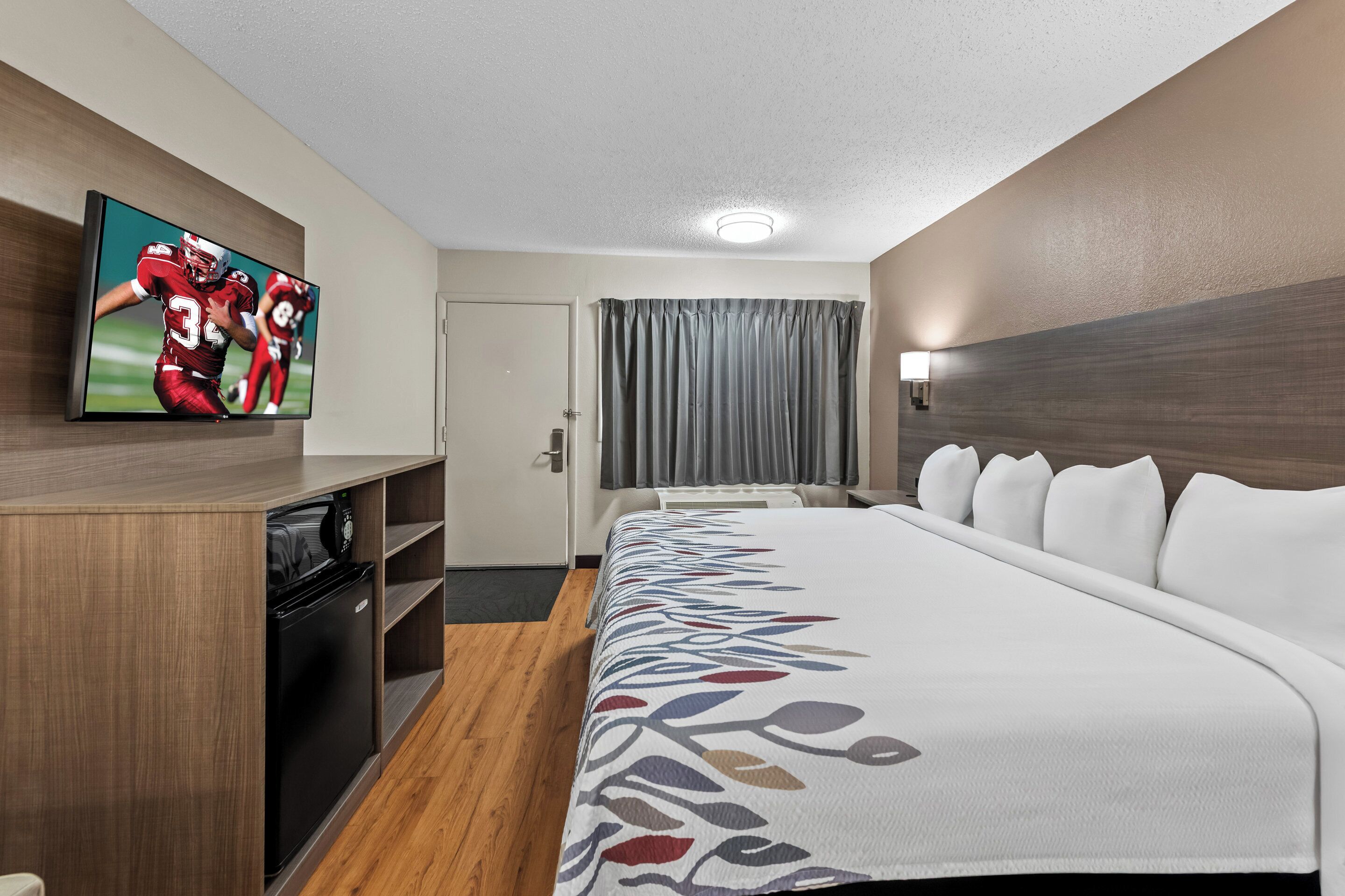 A hotel room with a bed, a television on the wall, a desk with a computer, and a door.