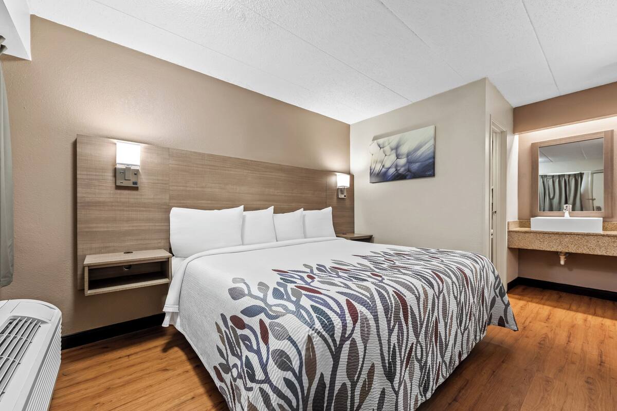 Quarto de hotel com uma cama grande, mesa de cabeceira, luminária na parede, quadro, banheiro com pia e espelho.