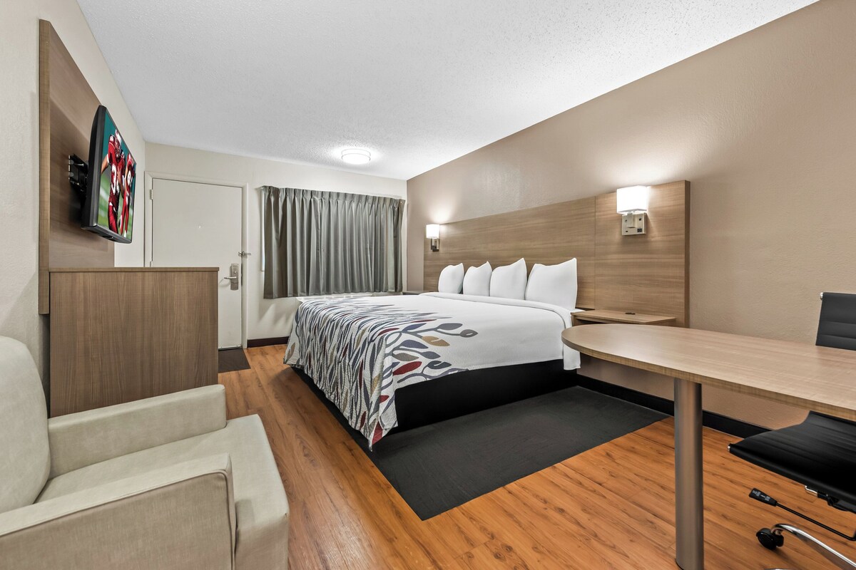 A hotel room with a bed, a desk, a chair, a flat-screen TV, and a wall-mounted light.