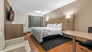 A hotel room with a bed, a desk, a chair, a flat-screen TV, and a wall-mounted light.