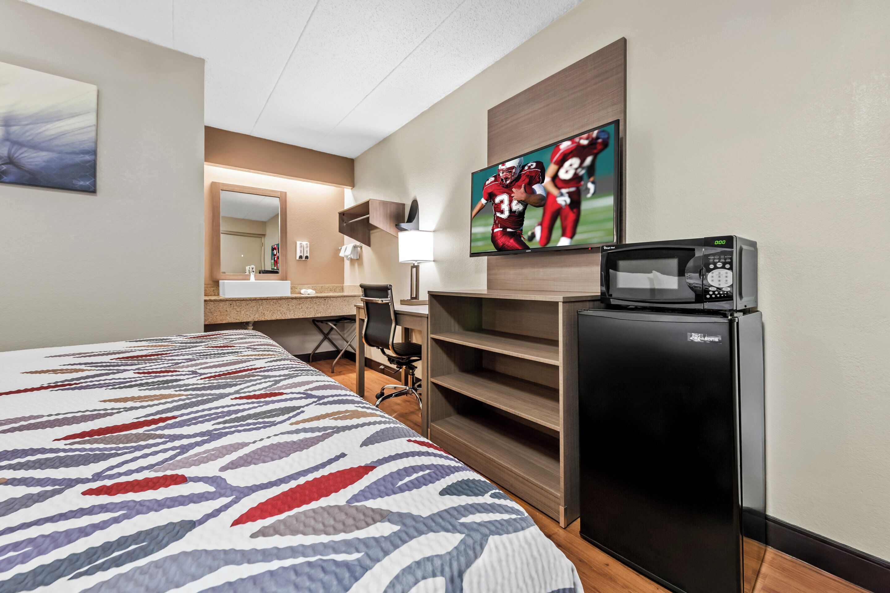 A hotel room with a bed, a TV, a refrigerator, a desk, and a chair.