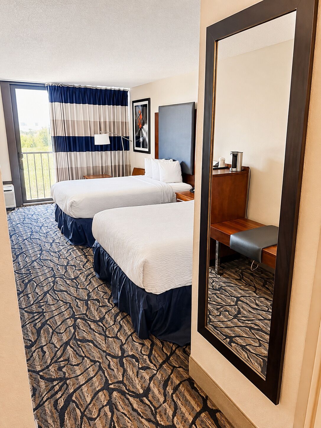 A hotel room with two beds, a large mirror, a desk, and a view of the outdoors.