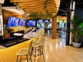 A modern bar area with multiple screens, wicker chairs, and a bar counter.