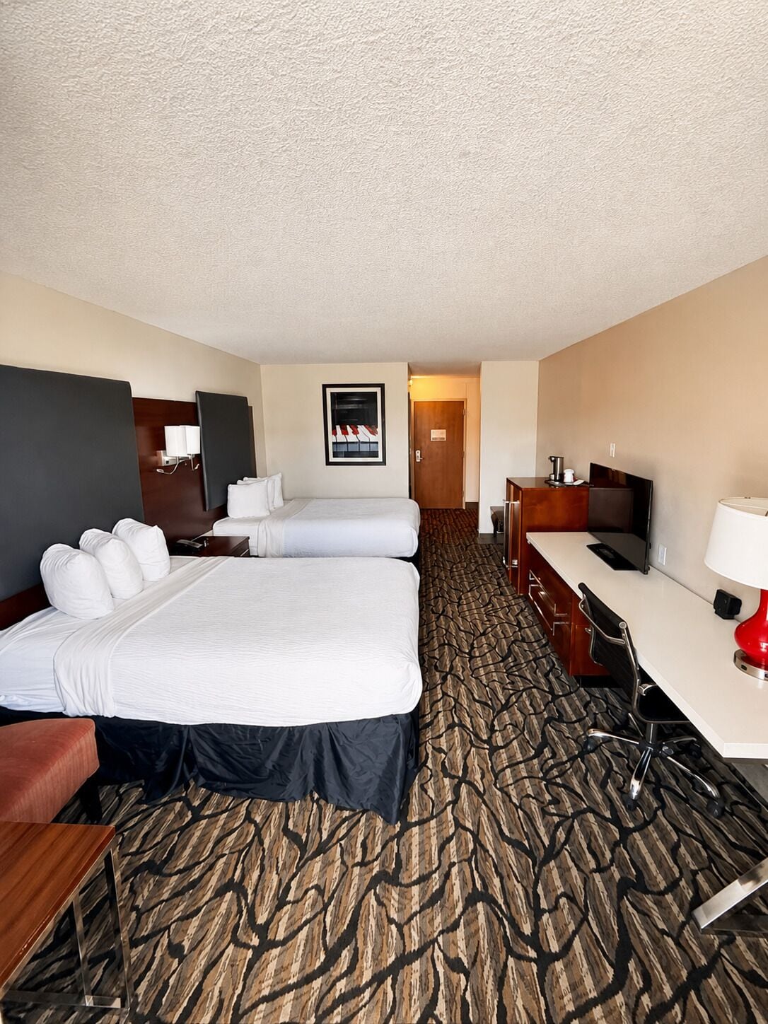A hotel room with a large bed, a desk, a chair, a television, and a door leading to another room.
