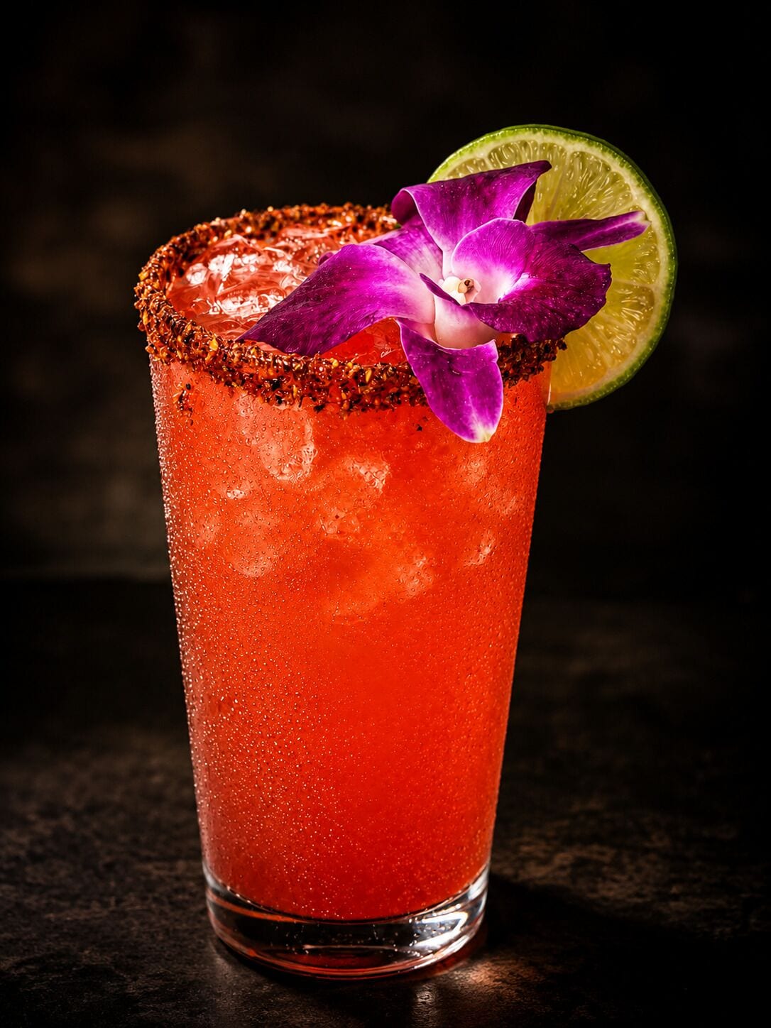 A tall glass with a salted rim, containing a red beverage, garnished with a purple flower and a lime slice.