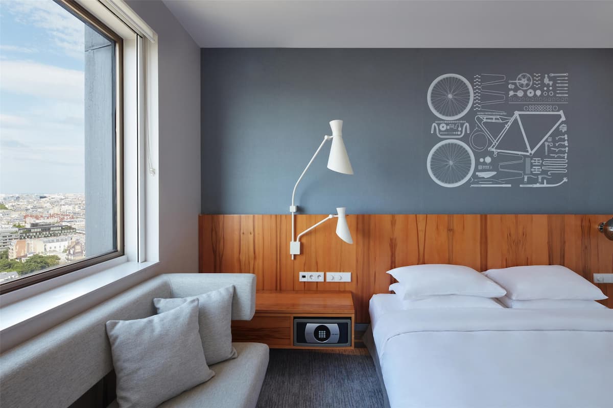 Hotellihuone, jossa on sänky, sohva, työpöytä, lamppu ja seinä, josta avautuu näkymä kaupunkimaisemaan.