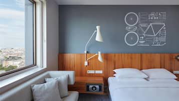 Ett hotellrum med en säng, en soffa, ett skrivbord, en lampa och en vägg med utsikt över stadsbilden.