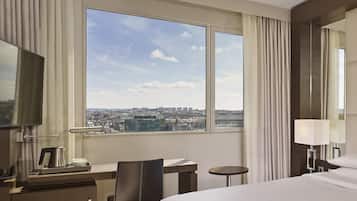 A hotel room with a bed, a desk with a coffee maker, a chair, a small table, and a large window offering a city view.