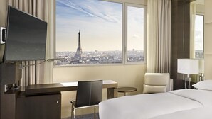 A hotel room with a bed, a desk, a chair, a TV, and a large window with a view of the Eiffel Tower. - Hyatt Regency Paris Etoile (Paris)
