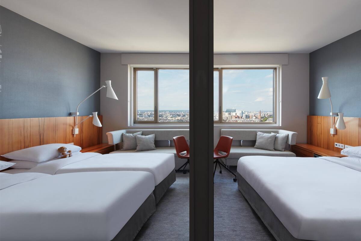 Hotellihuone, jossa on kaksi sänkyä, sohva, tuoli ja suuri ikkuna, josta avautuu näkymä kaupunkiin.