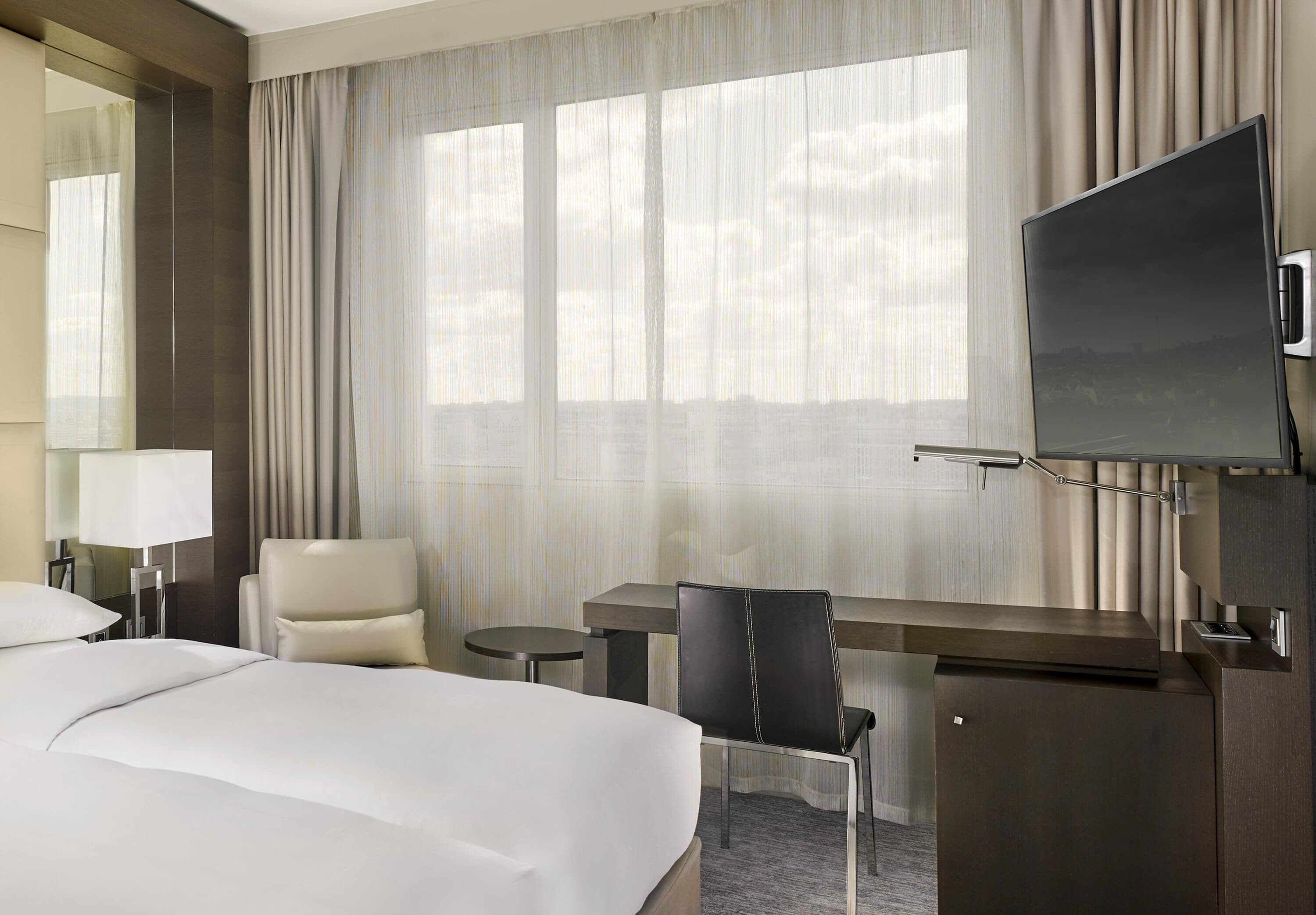 Una habitación de hotel con una cama grande, un escritorio con silla, televisor y ventana con cortinas.