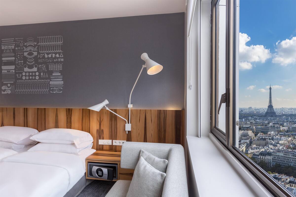 Moderni hotellihuone, jossa on sänky, harmaa sohva, lamppuvarustettu työpöytä ja näkymä kaupunkiin, josta näkyy Eiffel-torni.