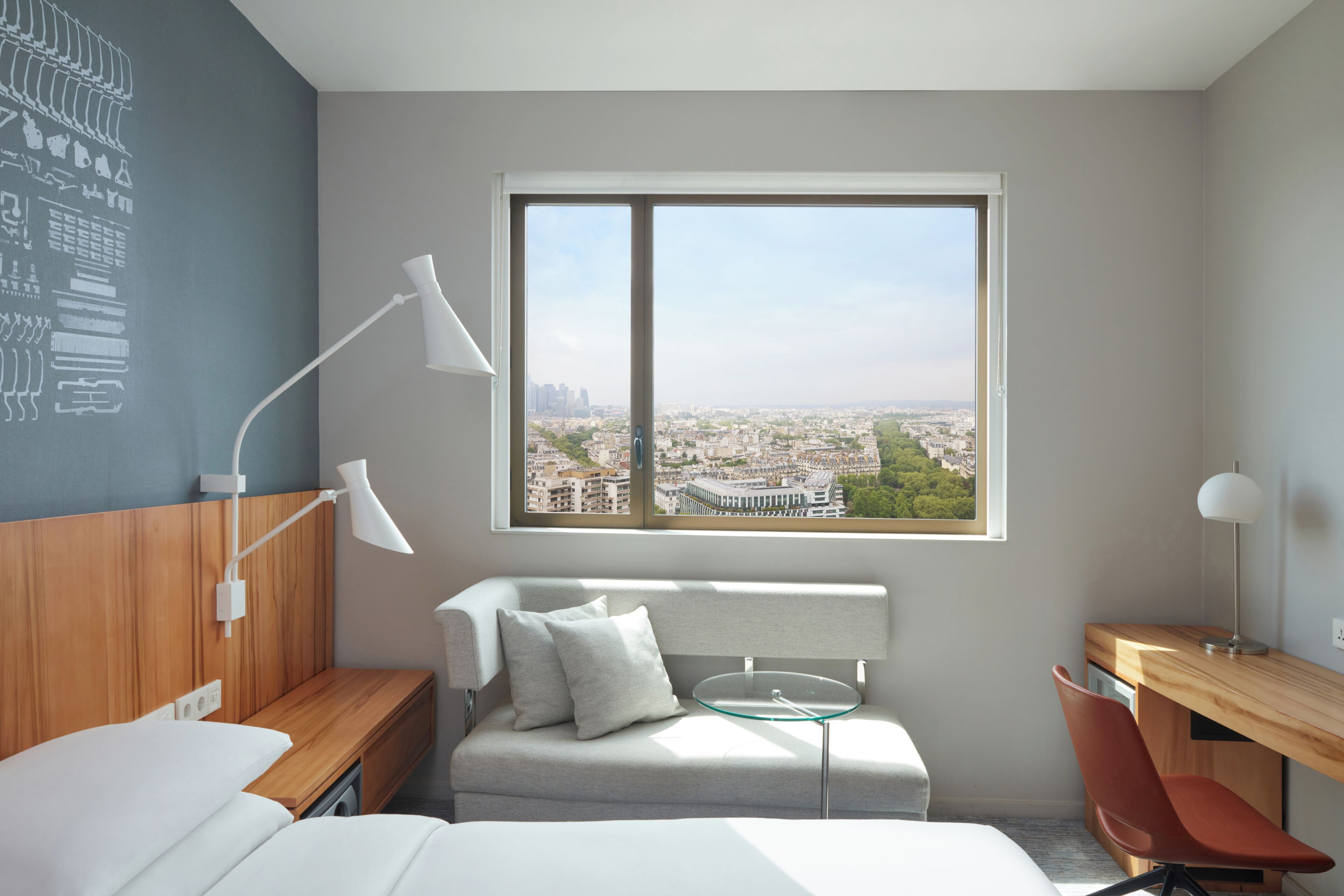 A modern hotel room with a bed, a desk, a chair, a small table, and a large window offering a city view.