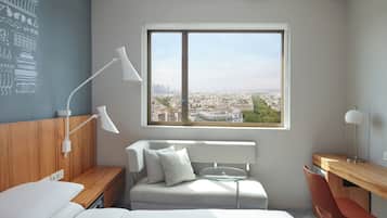 A modern hotel room with a bed, a desk, a chair, a small table, and a large window offering a city view.