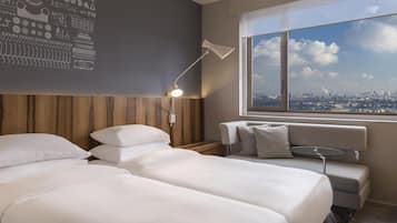 A hotel room with two beds, a nightstand, a lamp, a sofa, and a window with a city view.