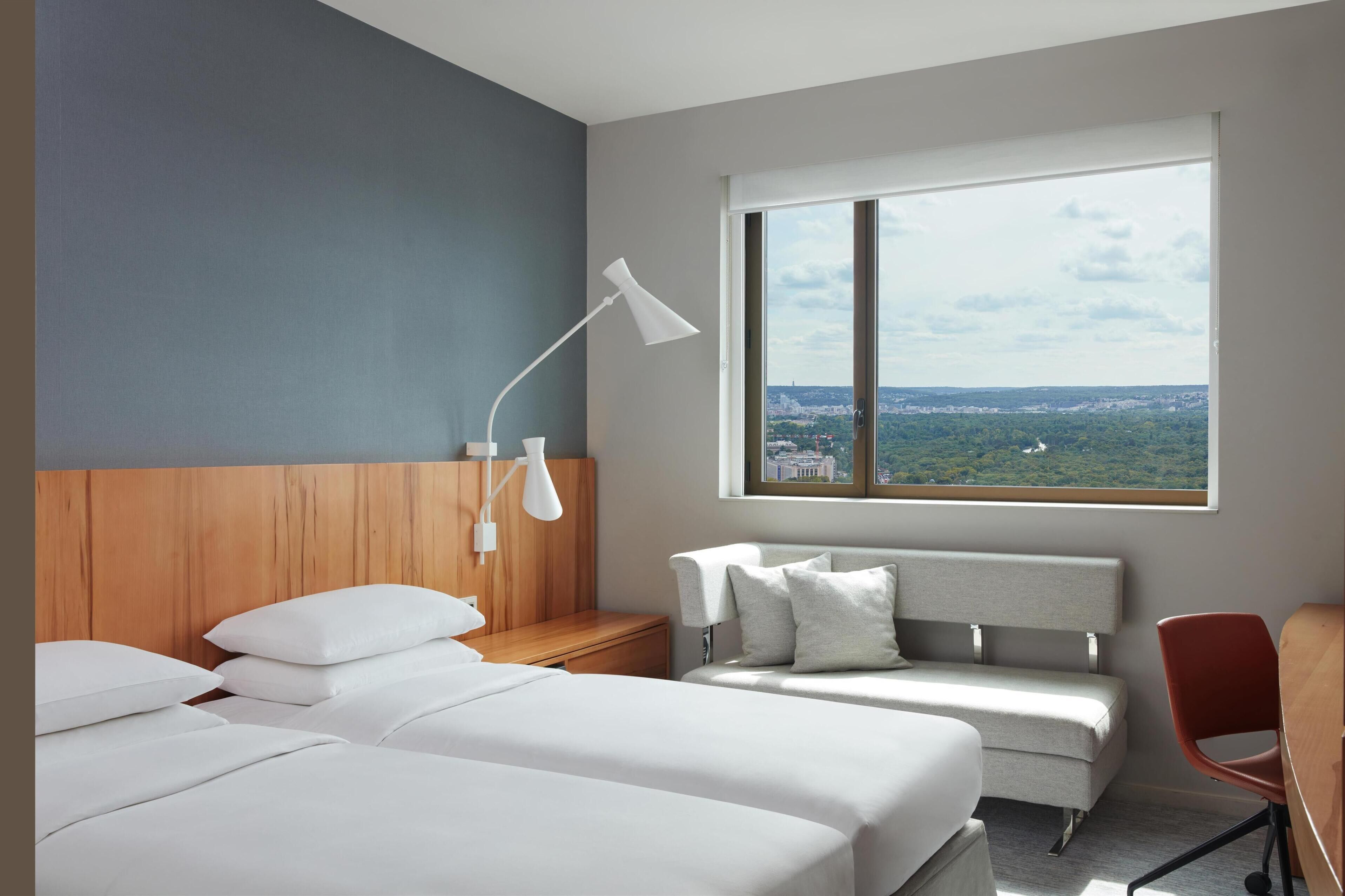 Habitación de hotel con dos camas, un escritorio, una silla, una mesita de noche, una lámpara y un amplio ventanal con vistas a la ciudad.