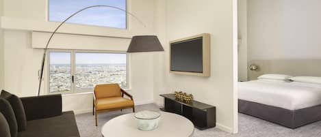 A modern hotel room with a large window offering a city view, a round table with a bowl, a sofa, a bed with white linens, and a mounted TV.