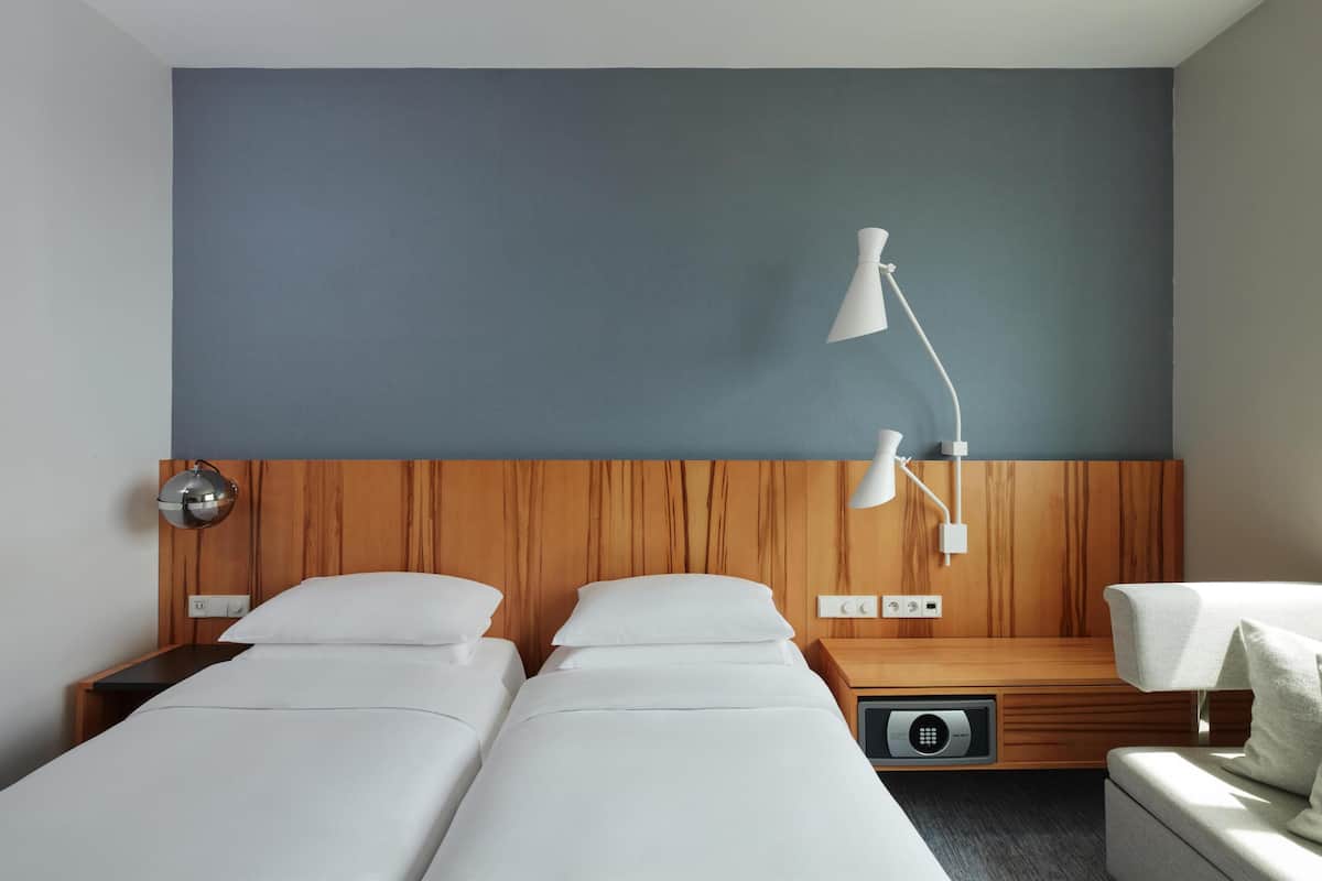 Hotellihuone, jossa on kaksi sänkyä, puinen pääty, yöpöytä lampun kanssa ja ohjauspaneeli.