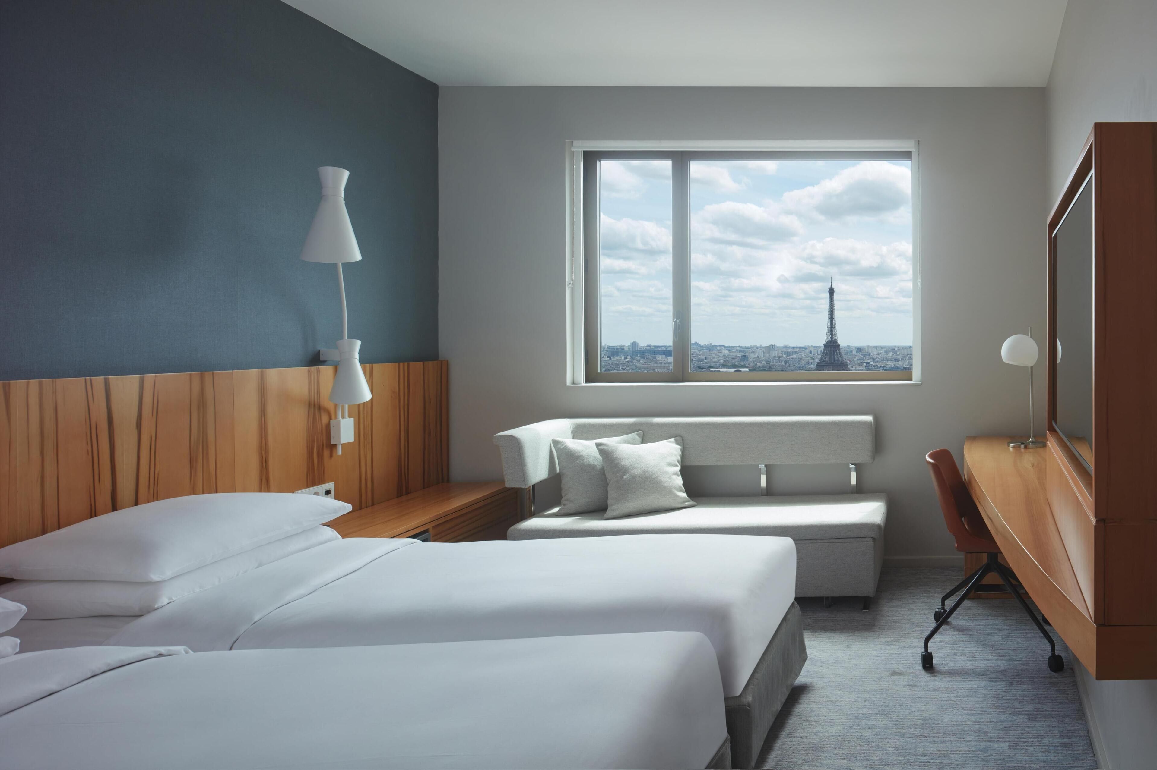 Habitación de hotel con dos camas, un escritorio, una silla, una lámpara y un amplio ventanal con vistas a la Torre Eiffel.