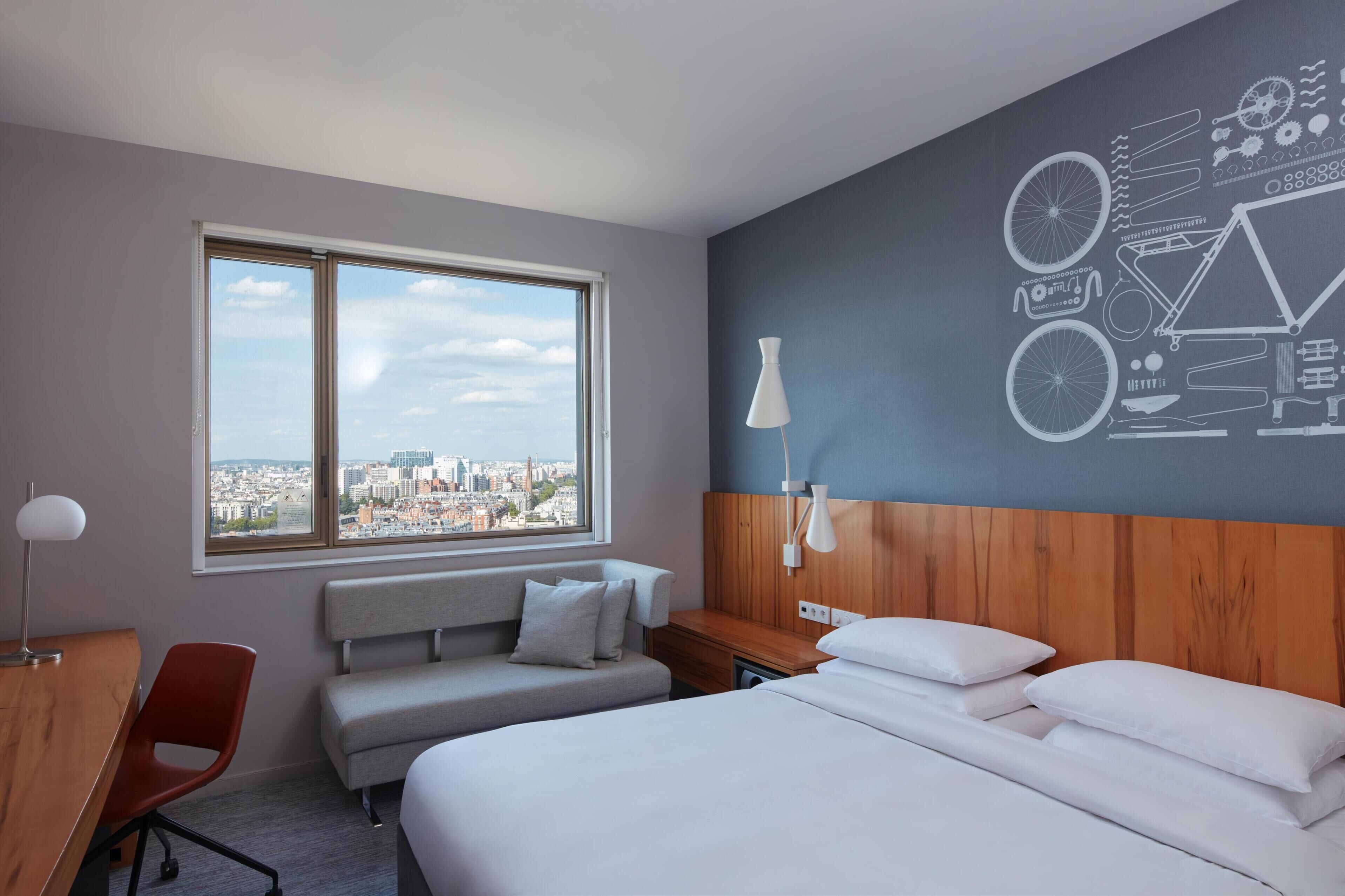 A hotel room with a large window, a bed, a desk, and a wall with a bicycle-themed design.