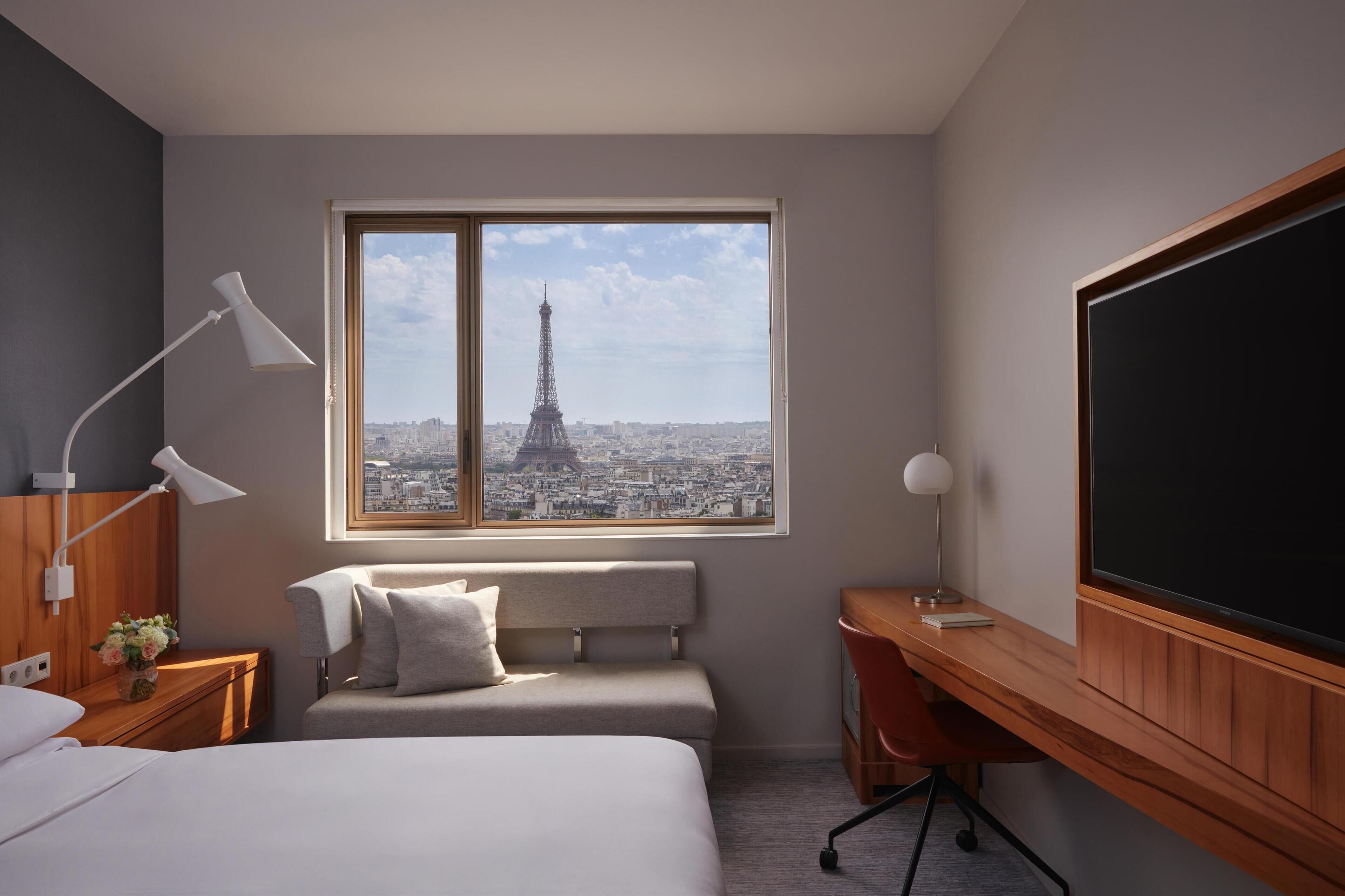 Quarto de hotel com cama, escrivaninha, cadeira, mesa de cabeceira, abajur e uma grande janela com vista para a Torre Eiffel.