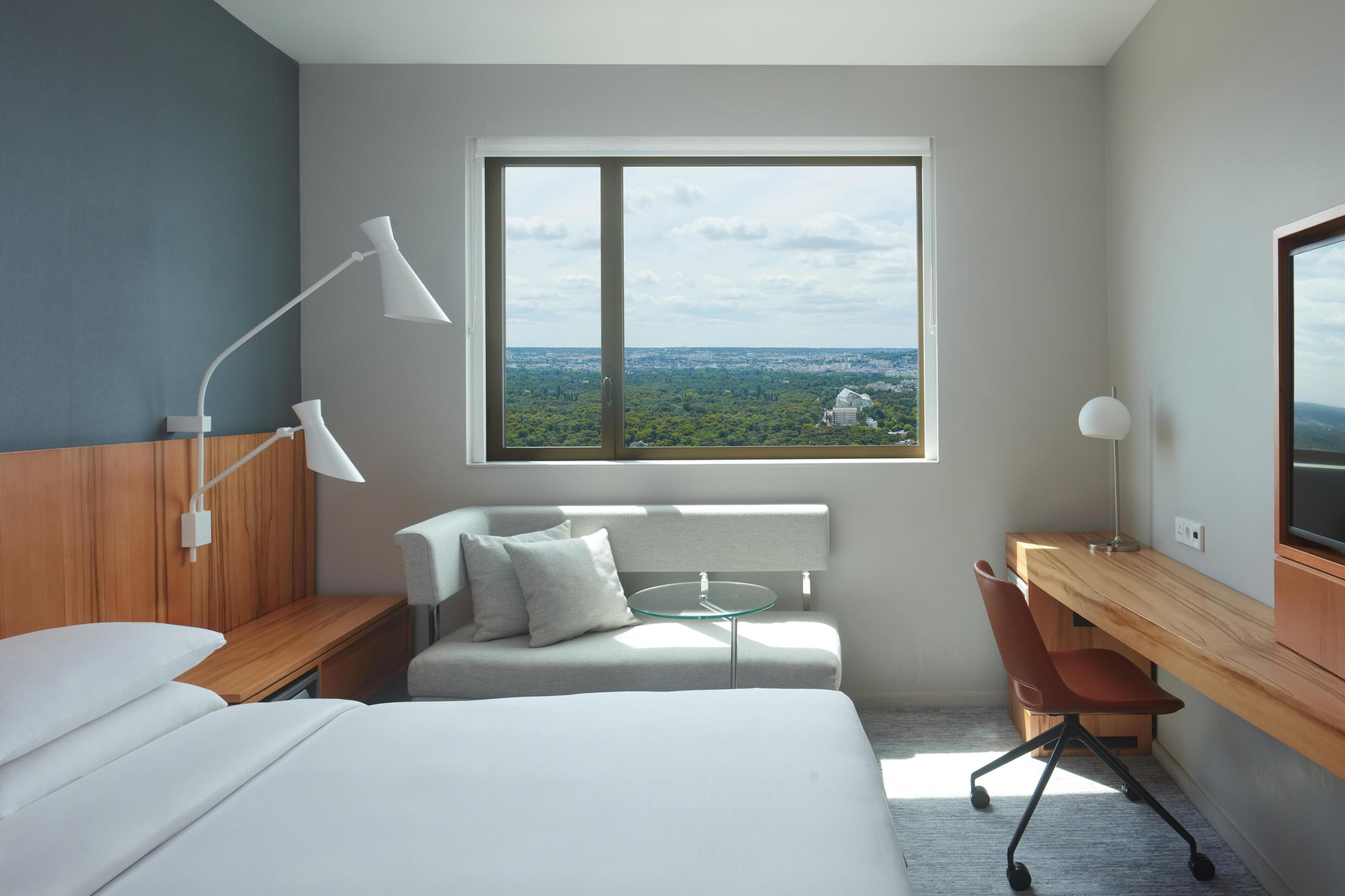 A modern hotel room with a bed, a desk, a chair, a small table, and a large window offering a city view.